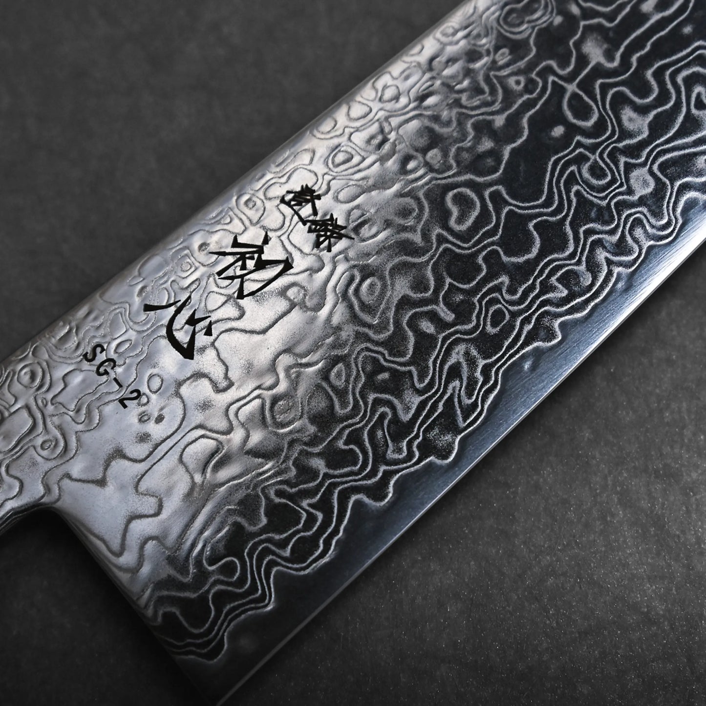 Close-up view of a Damascus steel kiritsuke Japanese chef knife blade featuring intricate, wavy patterns and Japanese characters etched on the upper part of the blade against a dark background.