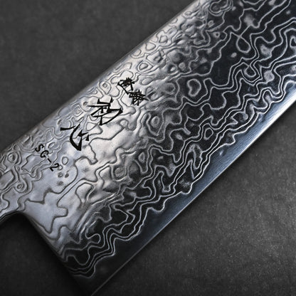 Close-up view of a Damascus steel kiritsuke Japanese chef knife blade featuring intricate, wavy patterns and Japanese characters etched on the upper part of the blade against a dark background.
