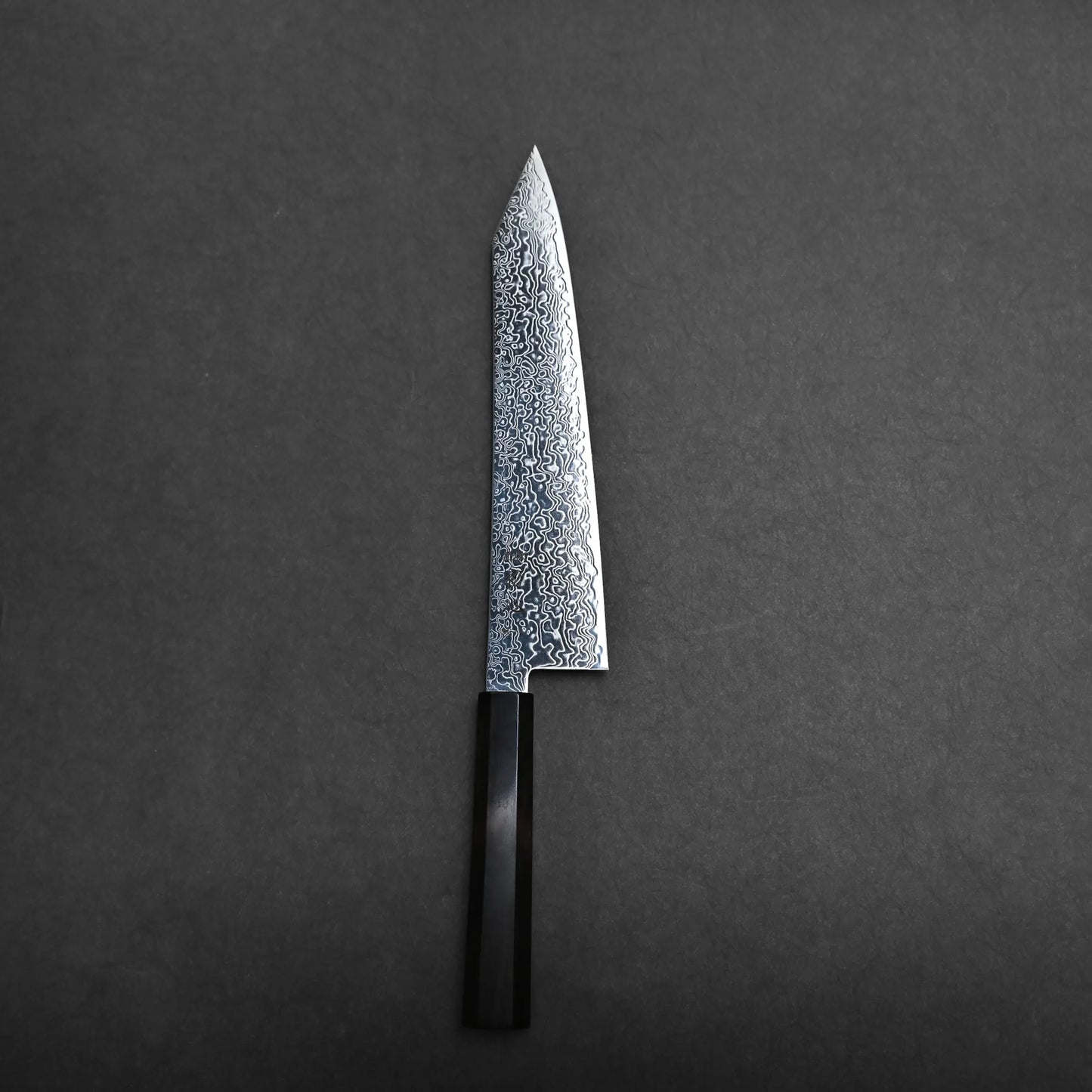 A Japanese-style kiritsuke gyuto knife with a patterned Damascus steel blade and a black handle is placed on a dark, textured surface.