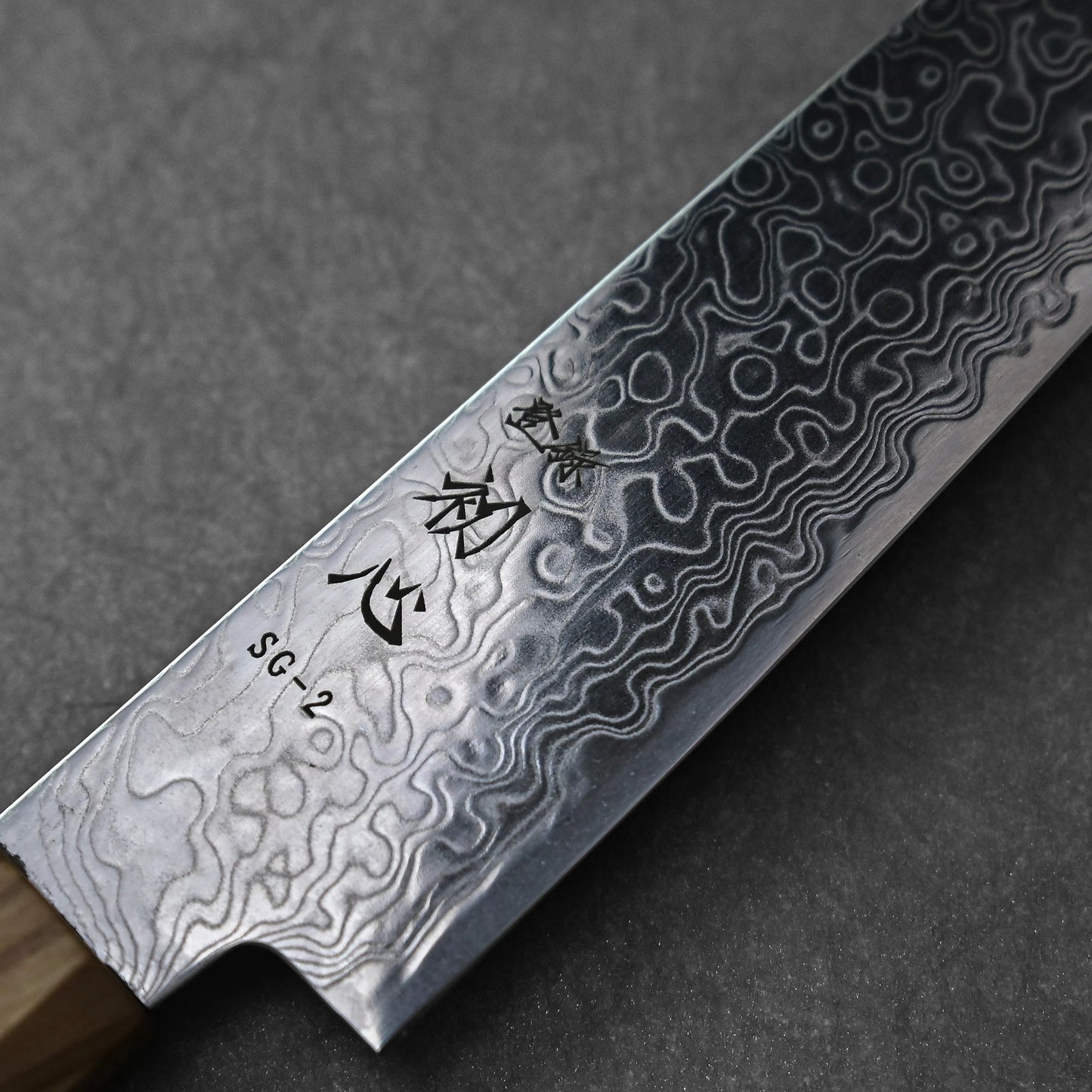 SG2 damascus petty knife with olive wood handle; photographed to highlight the blade’s intricate damascus pattern, engraved kanji near the SG2 steel marking, and the smooth grain of the olive wood handle. Set against a neutral background to emphasize the blade details.