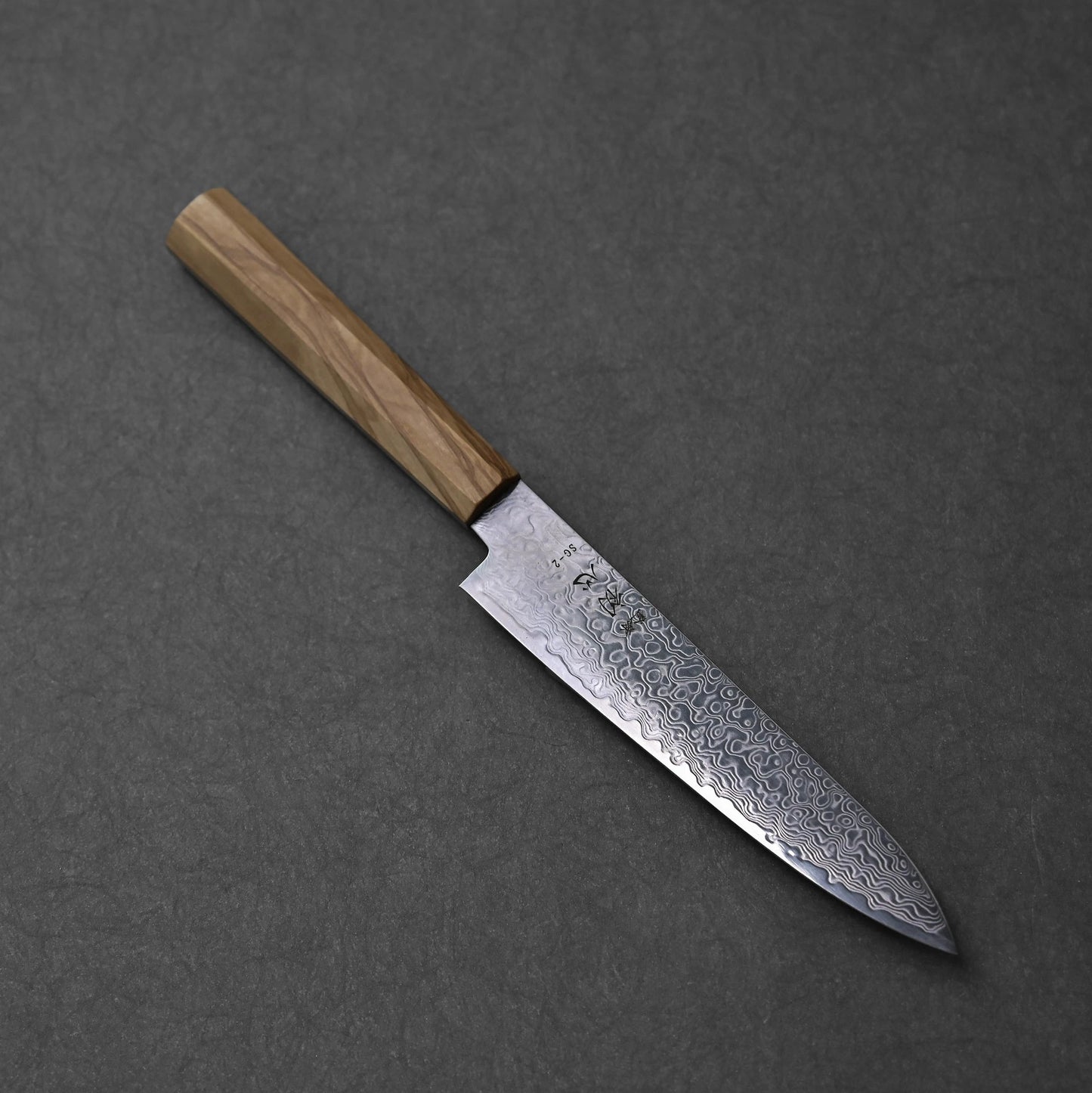 SG2 damascus petty knife with olive wood handle; photographed in angled orientation to show the front side of the blade, including the damascus pattern, SG2 marking, and wood grain.
