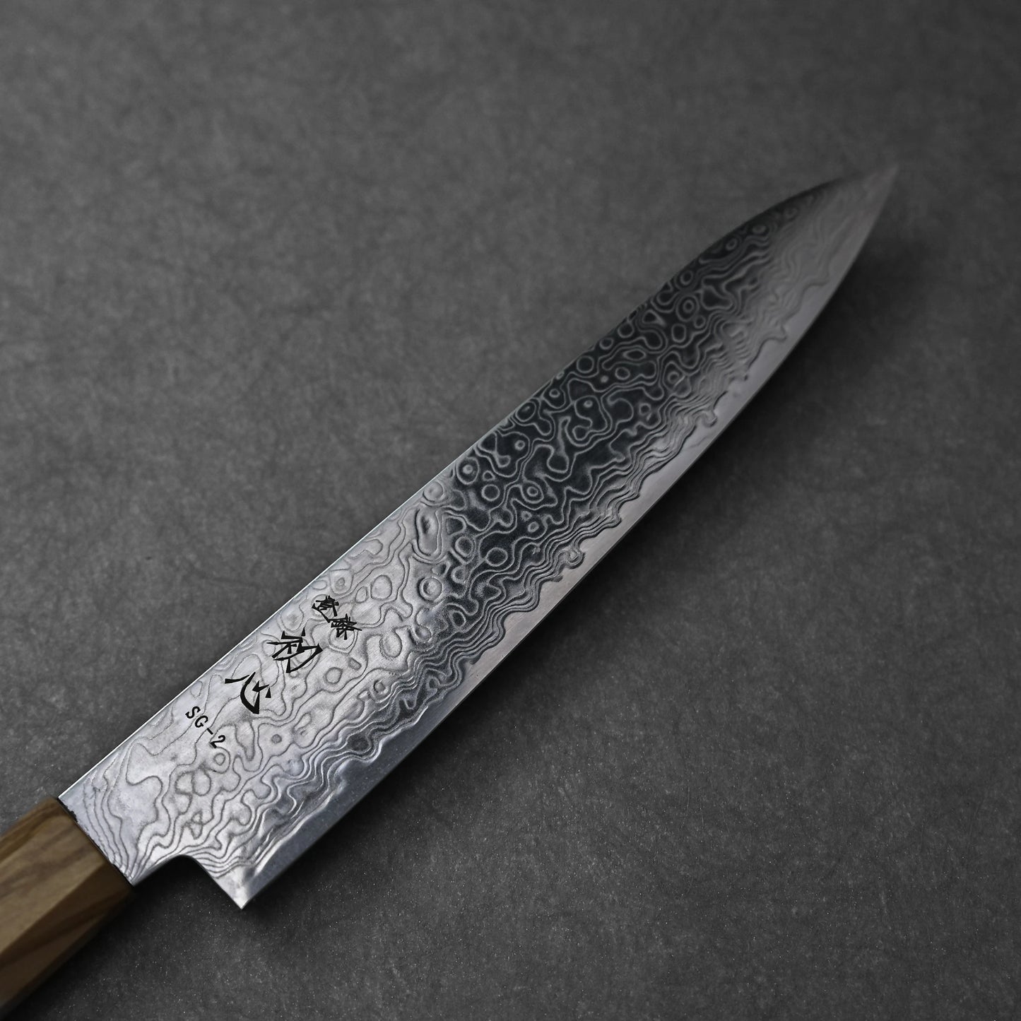 SG2 damascus petty knife with olive wood handle; close-up photograph highlighting the blade’s intricate damascus pattern, SG2 steel marking, engraved kanji, and the smooth grain of the olive wood handle near the ferrule.