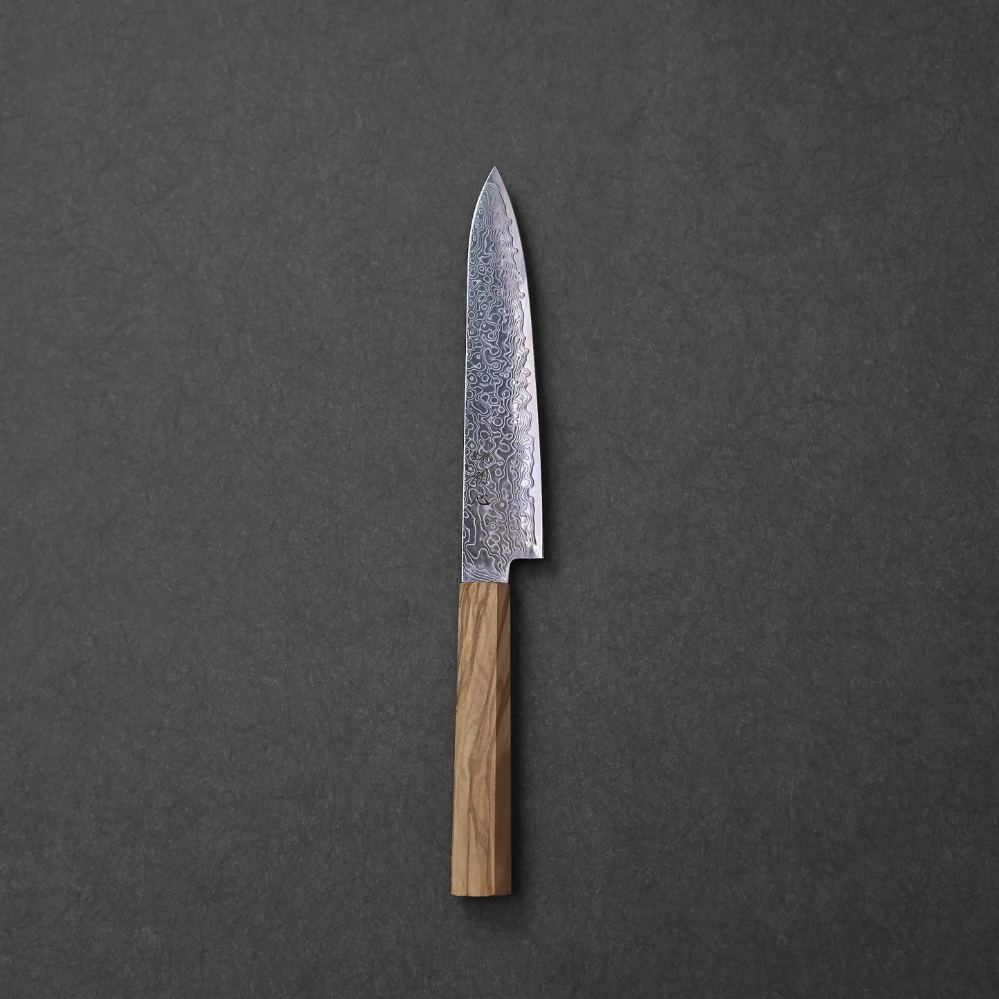 SG2 damascus petty knife with olive wood handle; photographed to highlight the blade’s flowing damascus pattern, compact shape, engraved kanji near the base, and the warm grain of the octagonal olive wood handle against a textured dark background.