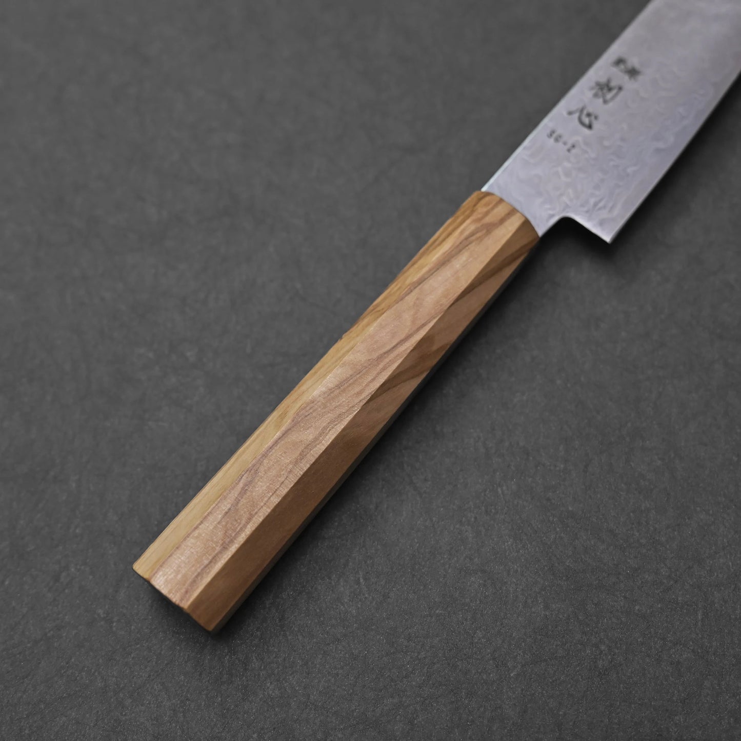 Close-up of the olive wood handle of an SG2 damascus petty knife; photographed to emphasize the faceted octagonal geometry, rich wood grain, and subtle transition to the blade with visible damascus texture.
