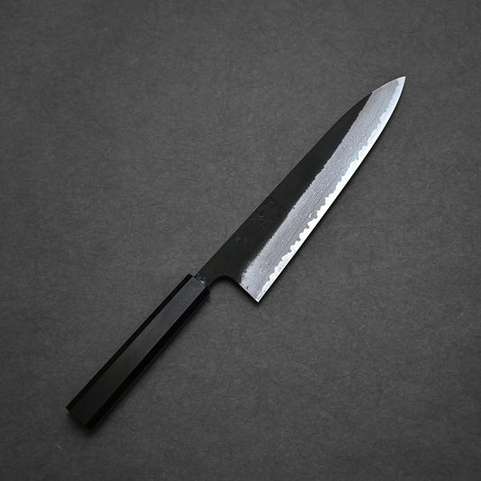 A Hatsukokoro Shinkiro kurouchi damascus AS gyuto 210mm  with a black handle and a silver, partially textured blade is placed on a dark gray surface, embodying the elegance of a kurouchi damascus gyuto.