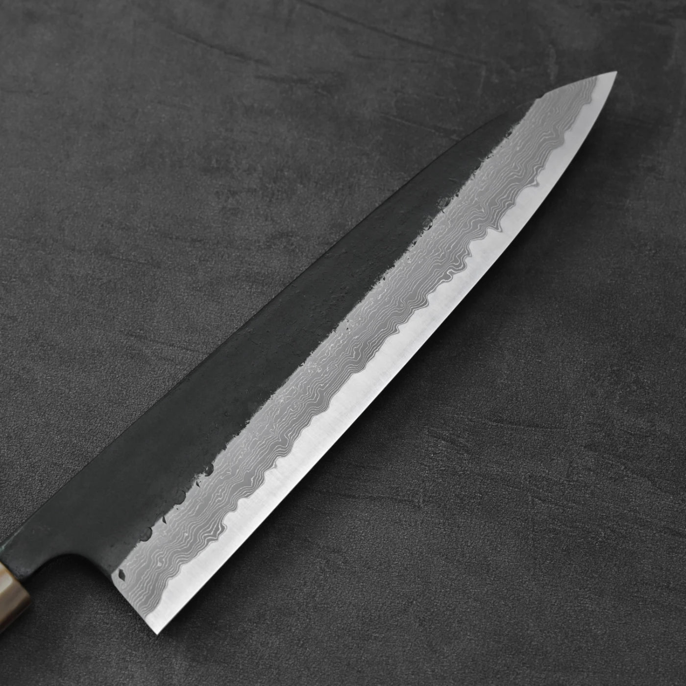 A gyuto with a kurouchi damascus finish is shown resting in a horizontal position.