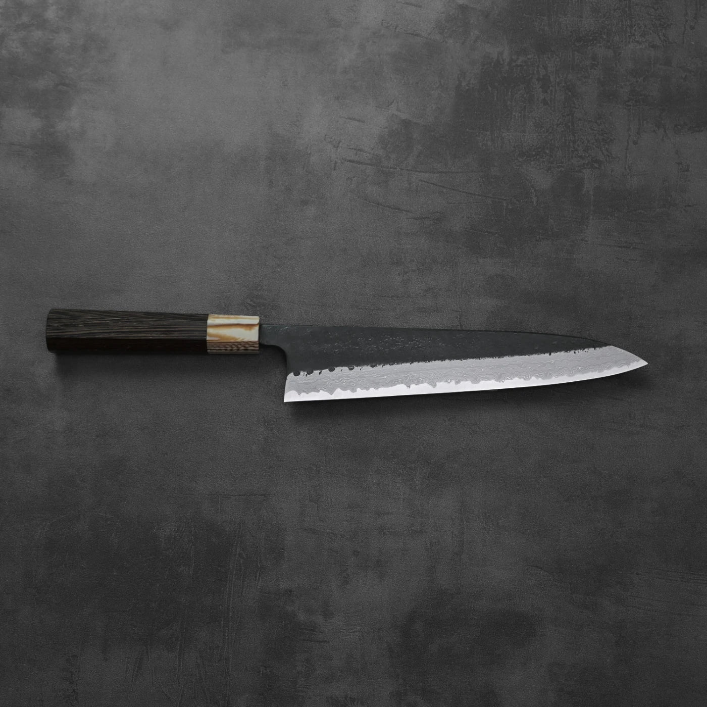 A gyuto knife with a kurouchi damascus finish rests, highlighting its blade and overall silhouette.