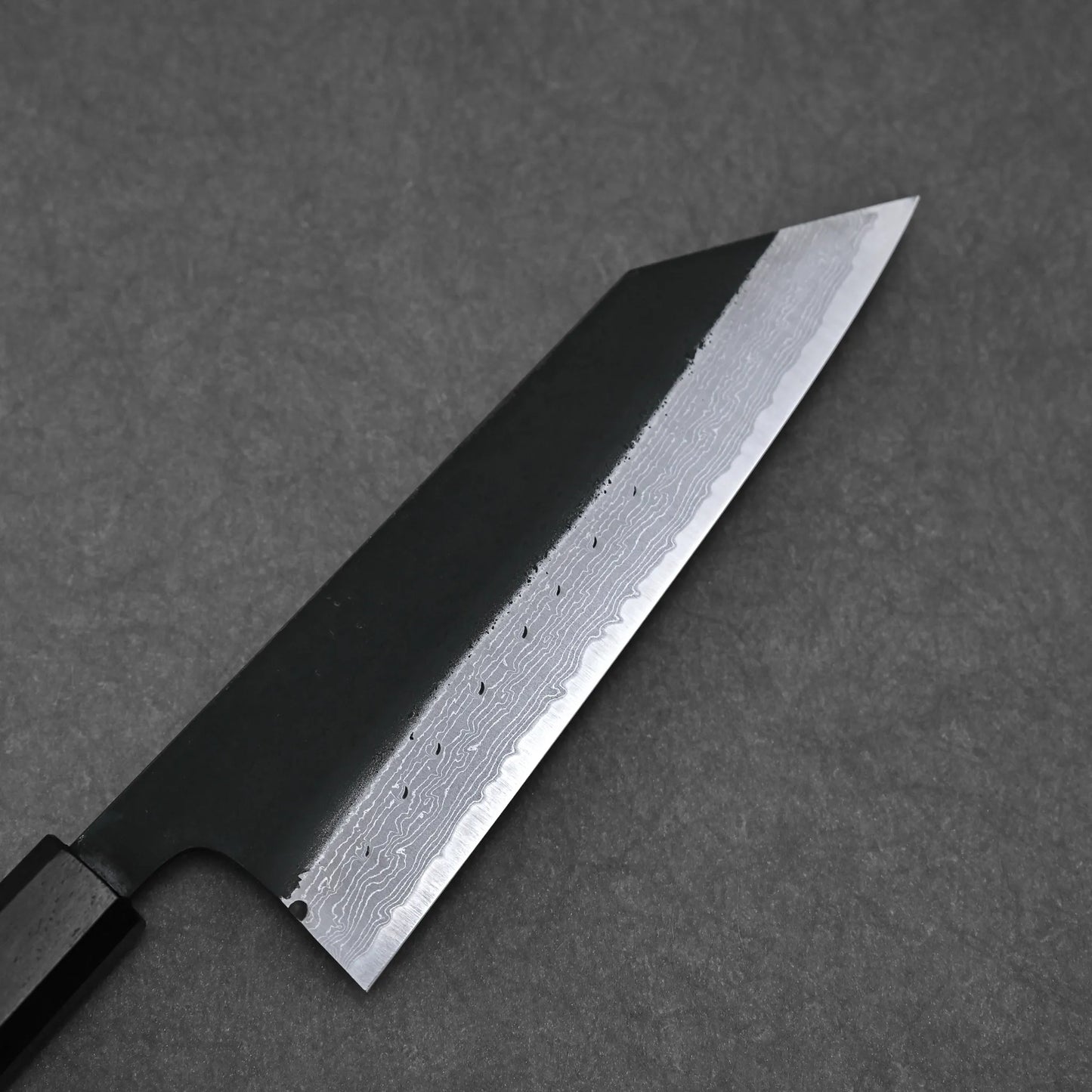 The Hatsukokoro Shinkiro kurouchi damascus AS bunka 170mm—featuring a dark handle and layered Damascus steel blade—rests on a textured gray surface, highlighting its exceptional Aogami Super craftsmanship.