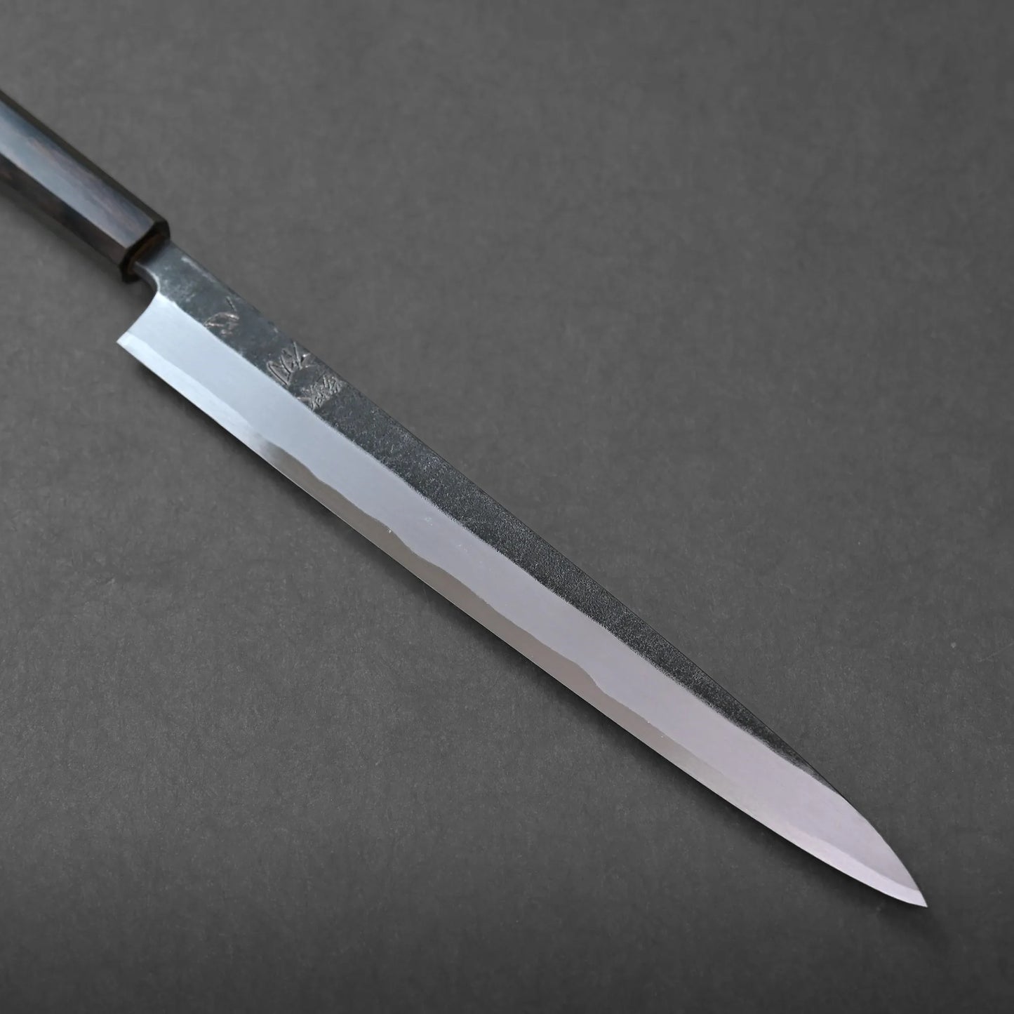 A long, narrow-bladed kitchen knife with a dark handle rests diagonally on a dark gray surface. The blade features a polished edge and a textured upper section, highlighting its craftsmanship.