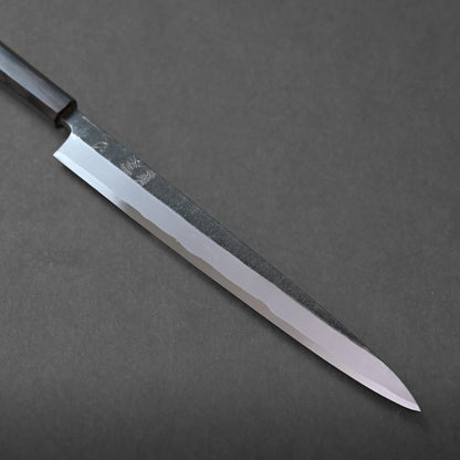 A long, narrow-bladed kitchen knife with a dark handle rests diagonally on a dark gray surface. The blade features a polished edge and a textured upper section, highlighting its craftsmanship.