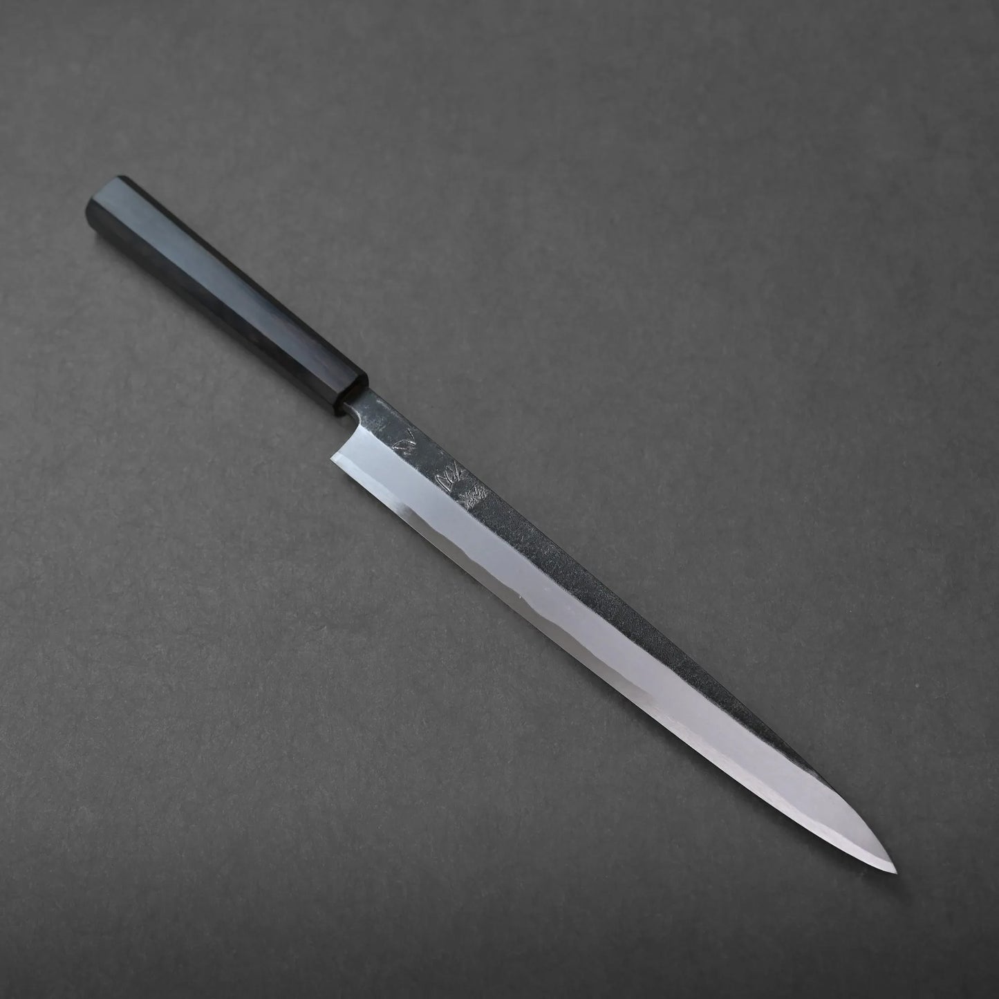 A Japanese-style kitchen knife with a long, narrow blade and a black handle rests on a dark gray surface, reflecting traditional craftsmanship.