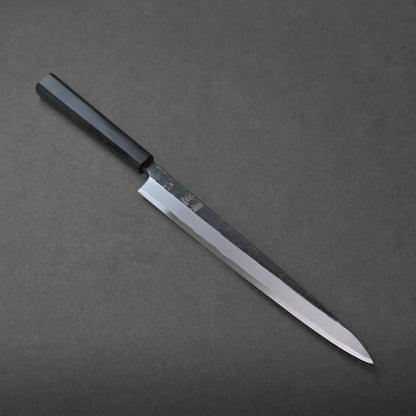 A Japanese-style kitchen knife with a long, narrow blade and a black handle rests on a dark gray surface, reflecting traditional craftsmanship.