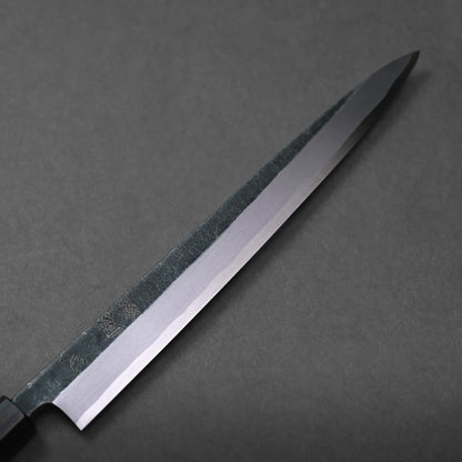 A close-up view of a long, narrow Japanese kitchen knife with a sleek blade and a dark handle, placed on a dark gray surface.