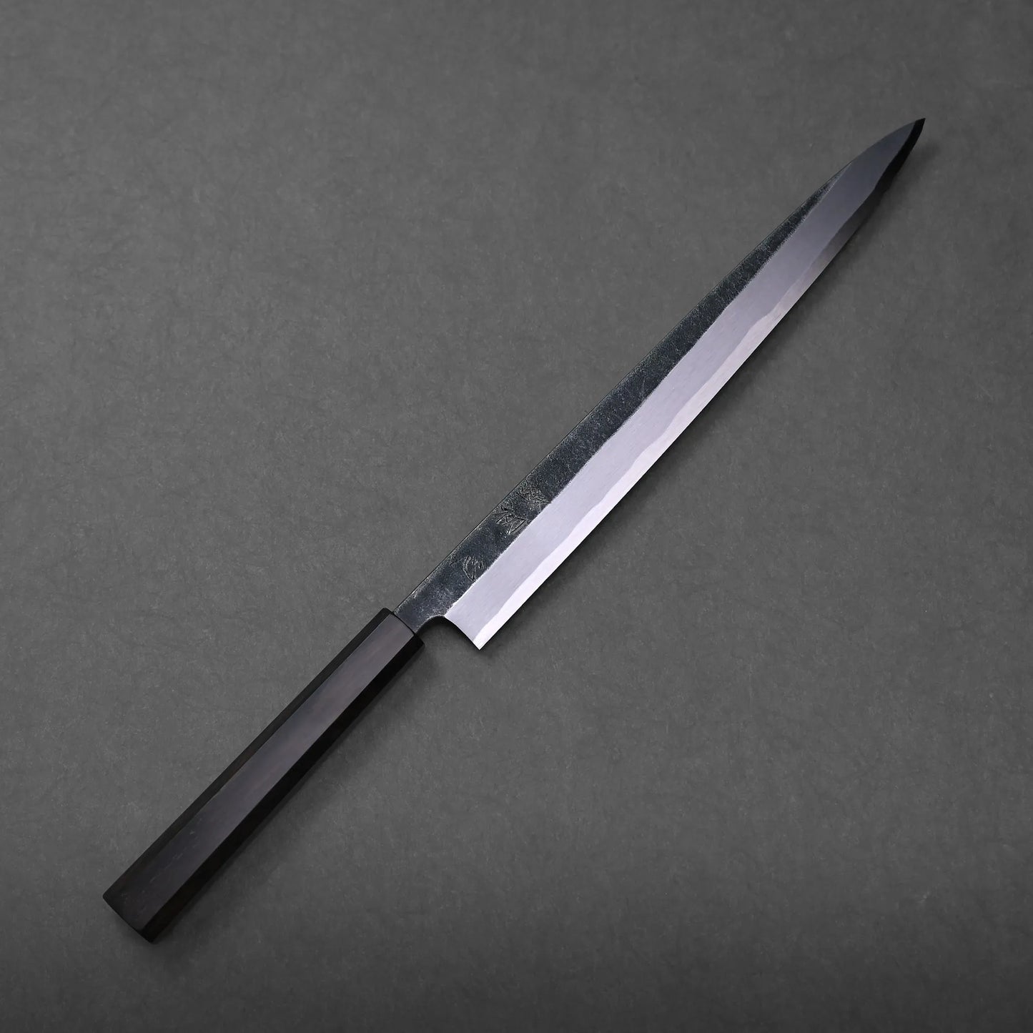 A Hatsukokoro Shirasagi kurouchi aogami#2 yanagiba 300mm with a dark handle rests on a plain, dark gray surface, its blade gleaming subtly.