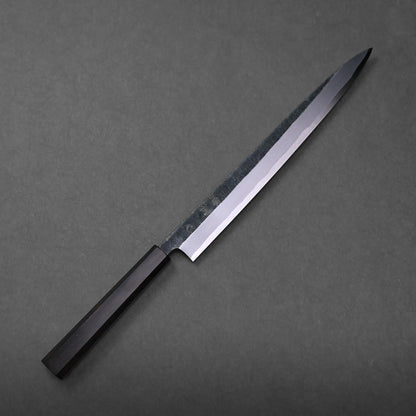 A Hatsukokoro Shirasagi kurouchi aogami#2 yanagiba 300mm with a dark handle rests on a plain, dark gray surface, its blade gleaming subtly.
