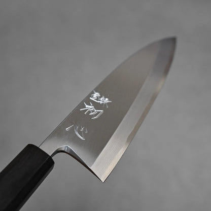 A close-up of the Hatsukokoro Shirasagi shirogami#2 deba 165mm, showing its shiny carbon steel blade with engraved Japanese characters and a black handle, set against a plain gray background.