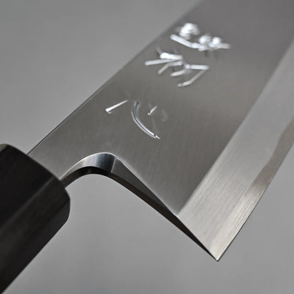 Close-up of the Hatsukokoro Shirasagi shirogami#2 deba 165mm, a Japanese fish knife made from Shirogami#2 carbon steel, displays engraved characters on its sharp blade and a dark handle against a plain grey background.
