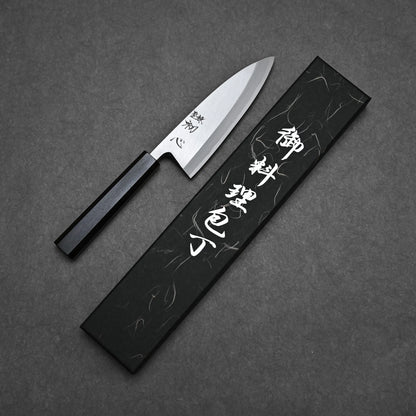 The Hatsukokoro Shirasagi Shirogami#2 Deba 165mm, featuring a black handle, lies beside a stylish black box with white Japanese characters on a dark textured surface, showcasing traditional craftsmanship and precise cutting performance.
