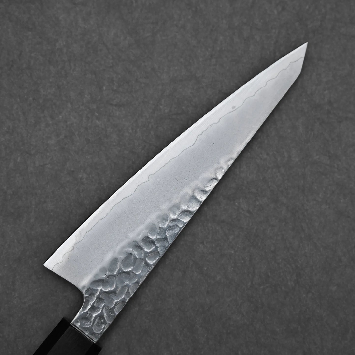 A close-up of the Hatsukokoro tsuchime ginsan honesuki 150mm chef knife, featuring a hammered tsuchime finish and wavy patterned blade, set against a dark gray textured background.