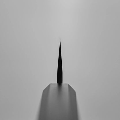 Close-up, black-and-white photo of the pointed tip of a Hatsukokoro tsuchime ginsan honesuki 150mm, shot from the handle end with sharp focus on the knife’s edge and a blurred background.