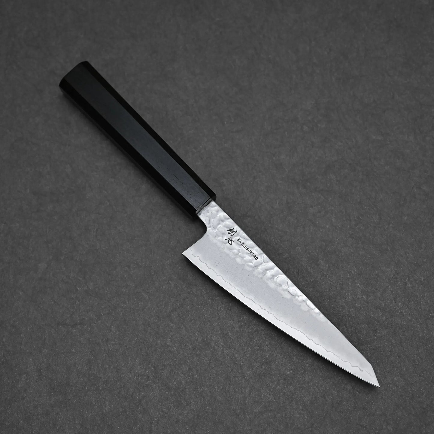 The Hatsukokoro tsuchime ginsan honesuki 150mm features a hammered finish, patterned blade, and black rectangular handle, displayed on a dark gray textured surface.