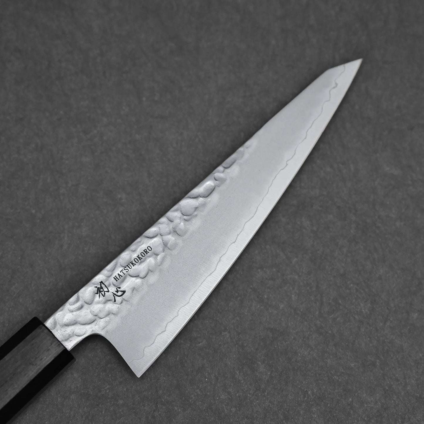 A close-up of the Hatsukokoro tsuchime ginsan honesuki 150mm displays its tsuchime finish, polished blade with engraved markings, and wooden handle as it rests elegantly on a dark textured surface.