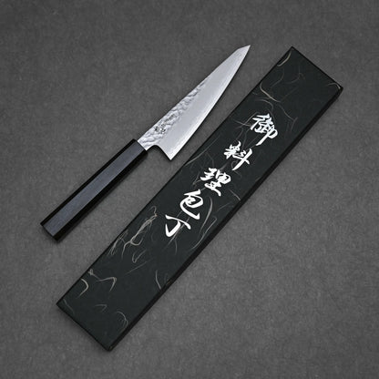 A Hatsukokoro tsuchime ginsan honesuki 150mm with a black handle sits next to a sleek black box featuring white Japanese characters, all displayed against a dark background.
