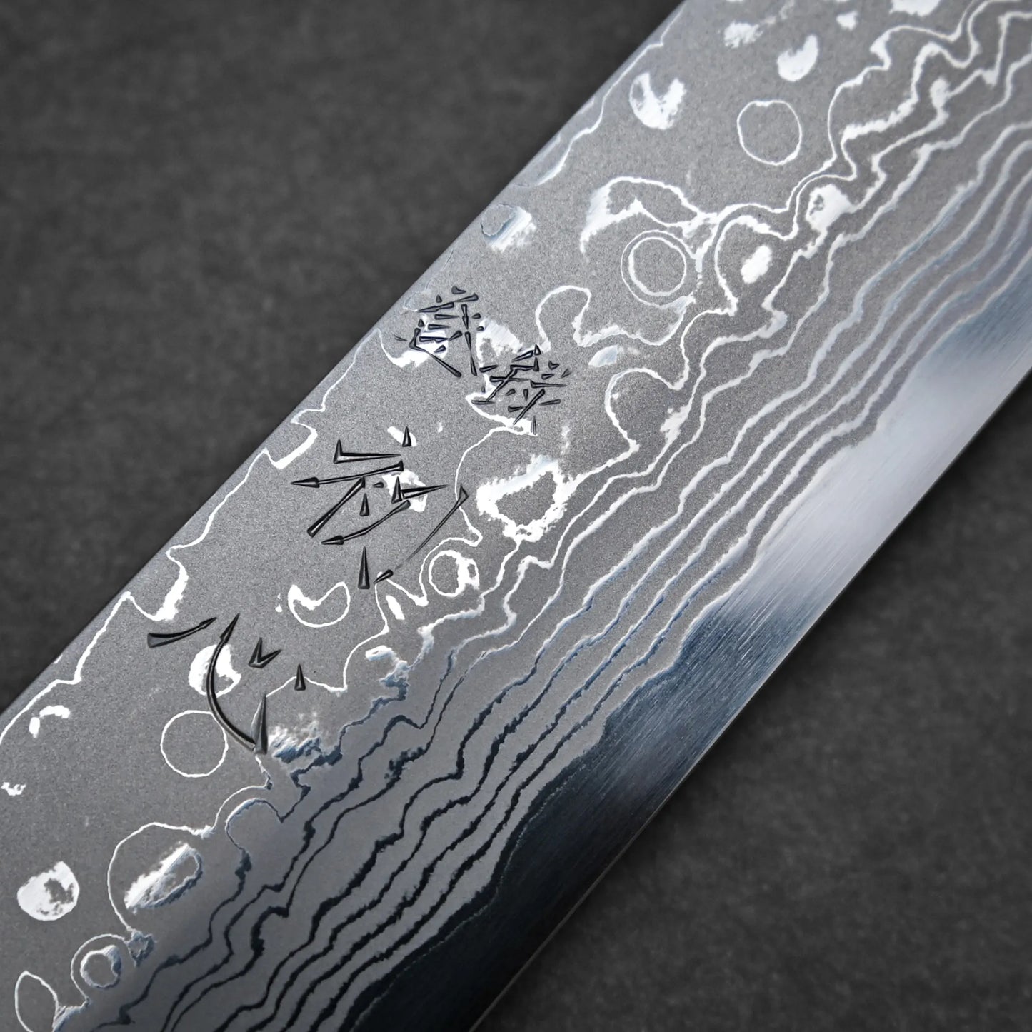 A close-up of a Damascus sashimi knife blade with visible wavy patterns and etched Japanese characters on its surface, set against a dark background.