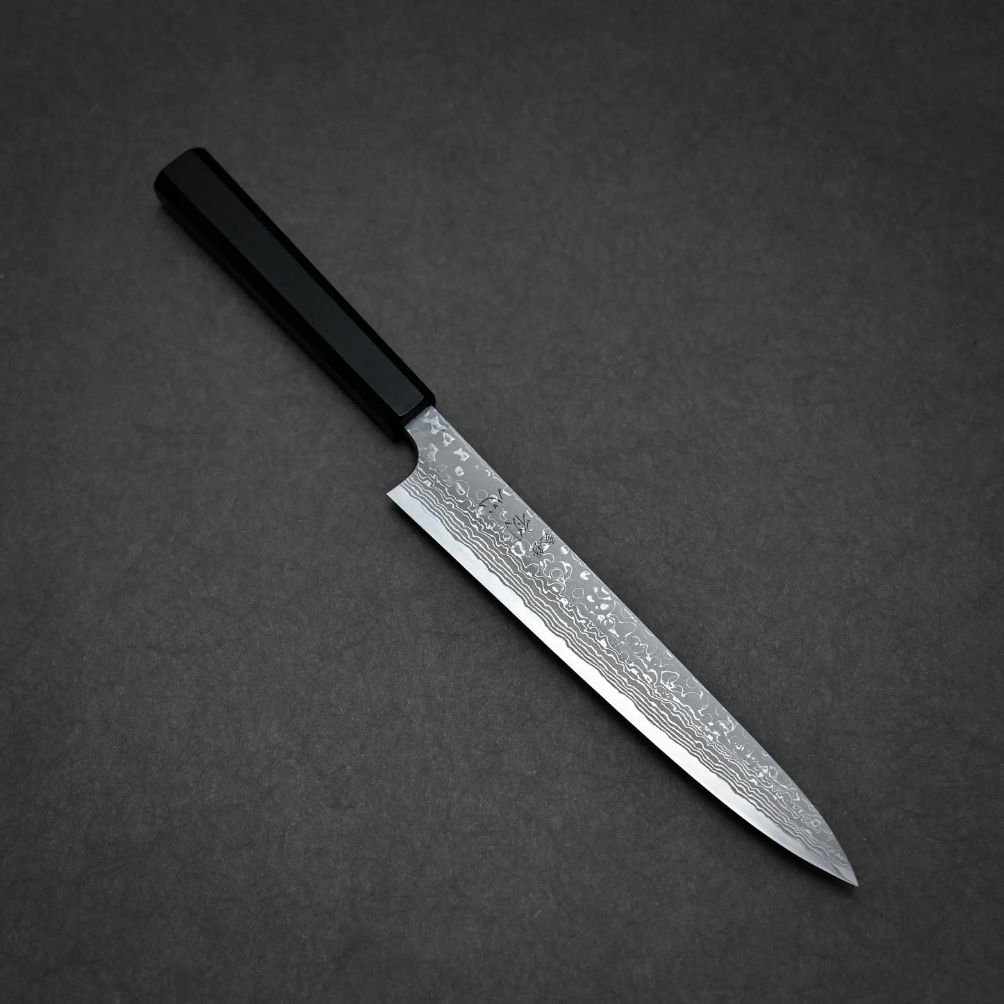 A Japanese-style damascus sashimi knife with a black handle and a patterned, narrow, pointed blade, placed on a dark grey surface.