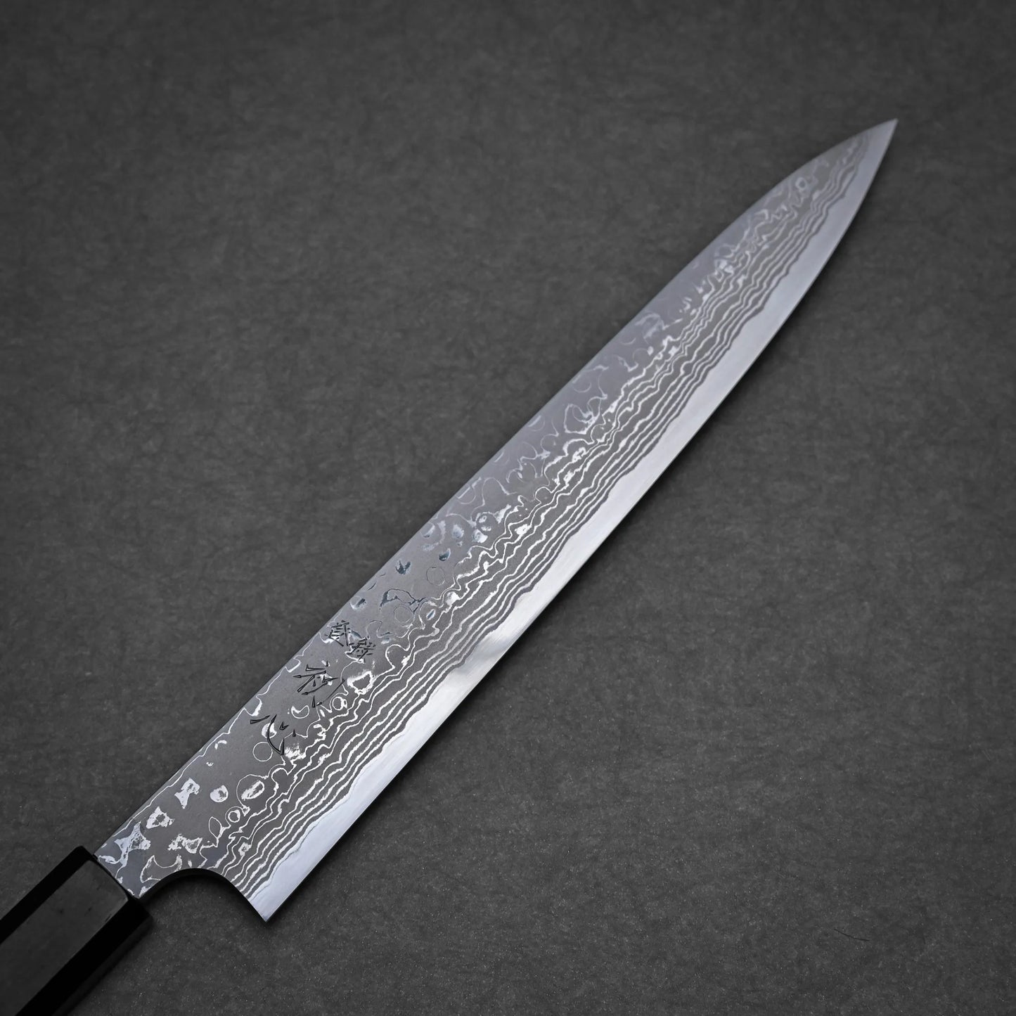 A close-up view of a long, narrow damascus sushi knife with a patterned Damascus steel blade and a dark handle, placed on a gray textured surface.