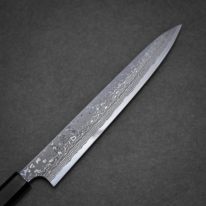 A close-up view of a long, narrow damascus sushi knife with a patterned Damascus steel blade and a dark handle, placed on a gray textured surface.