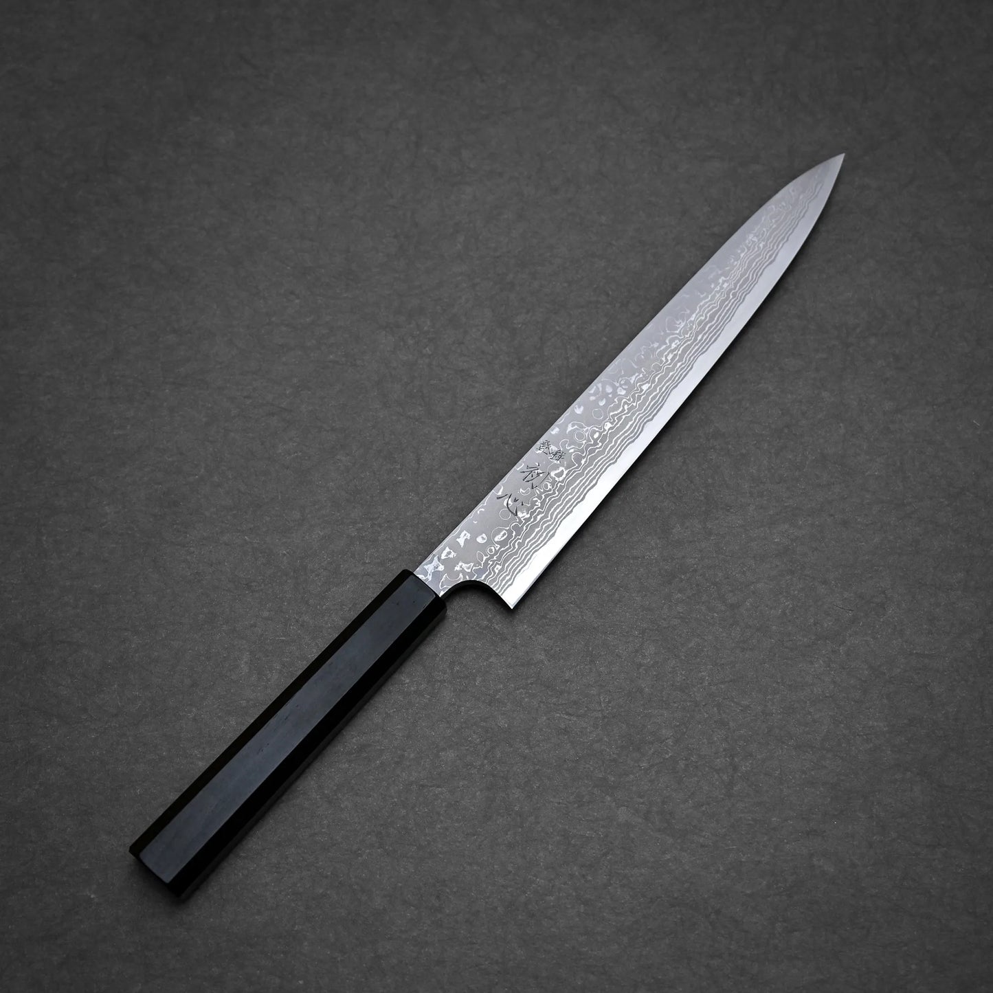 A Hatsukokoro x Yauji damascus aogami#2 yanagiba 240mm with a patterned blade and a black rectangular handle is placed on a dark gray textured surface.