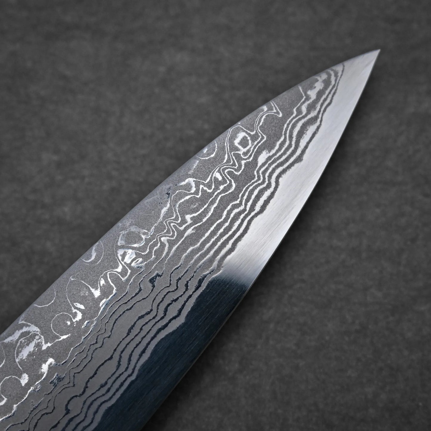 Close-up of a damascus sushi knife blade displaying distinctive wavy and patterned lines, with the tip pointing to the right against a dark background.
