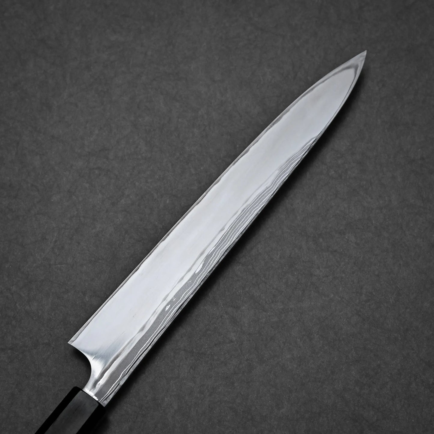 A close-up view of a Damascus yanagiba with a polished, reflective blade and dark handle, photographed on a dark, textured background.