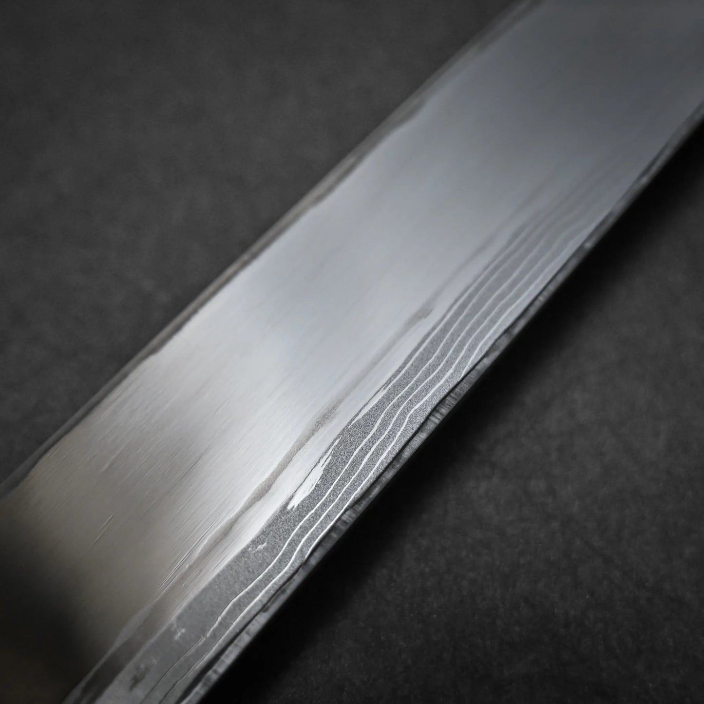 Close-up of a polished Damascus sushi knife blade with subtle wavy patterns around the uraoshi, set against a dark background.