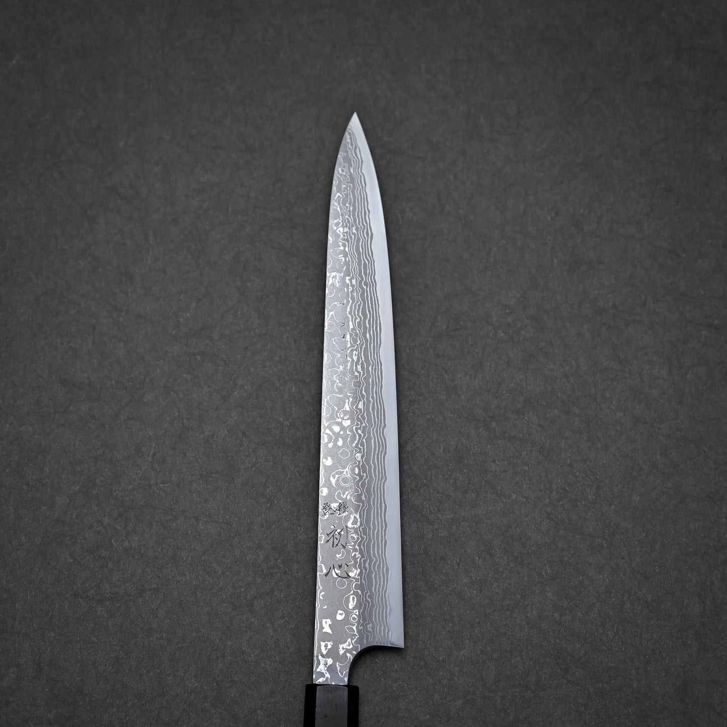 A close-up of a damascus yanagiba with a patterned Damascus steel blade, shown against a dark grey textured background.