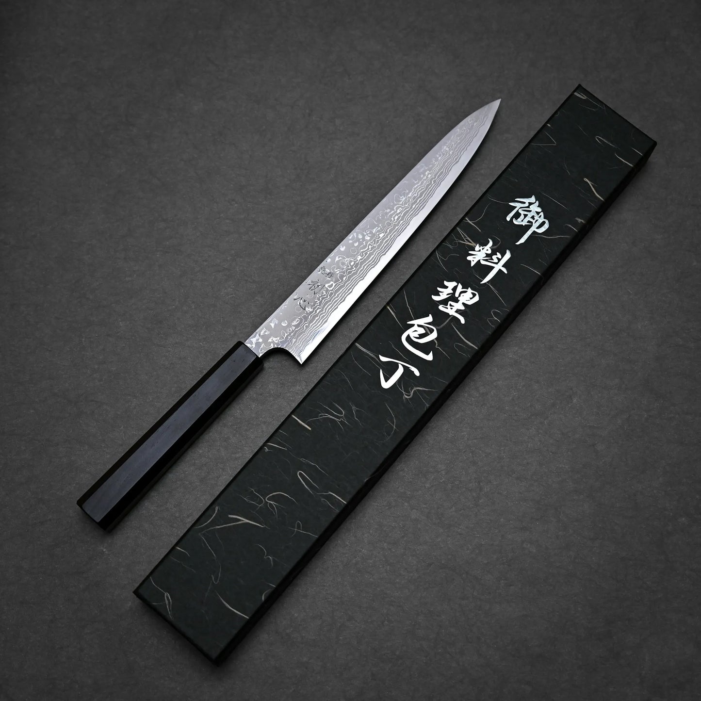A damascus sushi knife with a patterned blade and a black handle is placed next to a black box with silver Japanese characters on a dark textured surface.