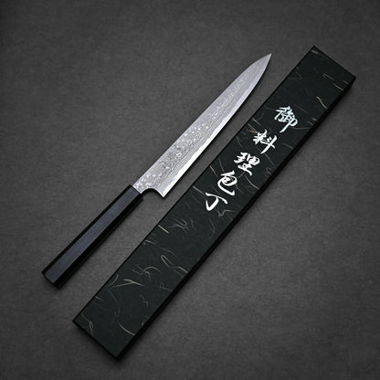A damascus sushi knife with a patterned blade and a black handle is placed next to a black box with silver Japanese characters on a dark textured surface.