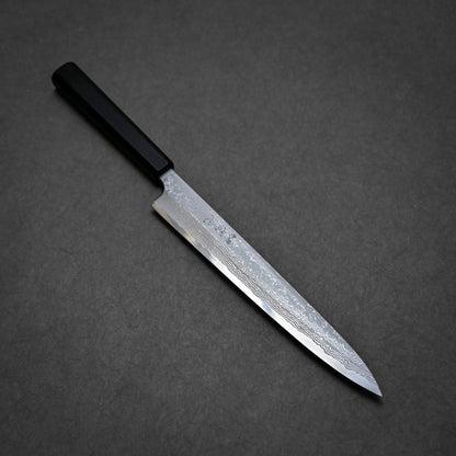 A damascus yanagiba with a dark rectangular handle and a long, patterned steel blade is placed on a dark gray surface.