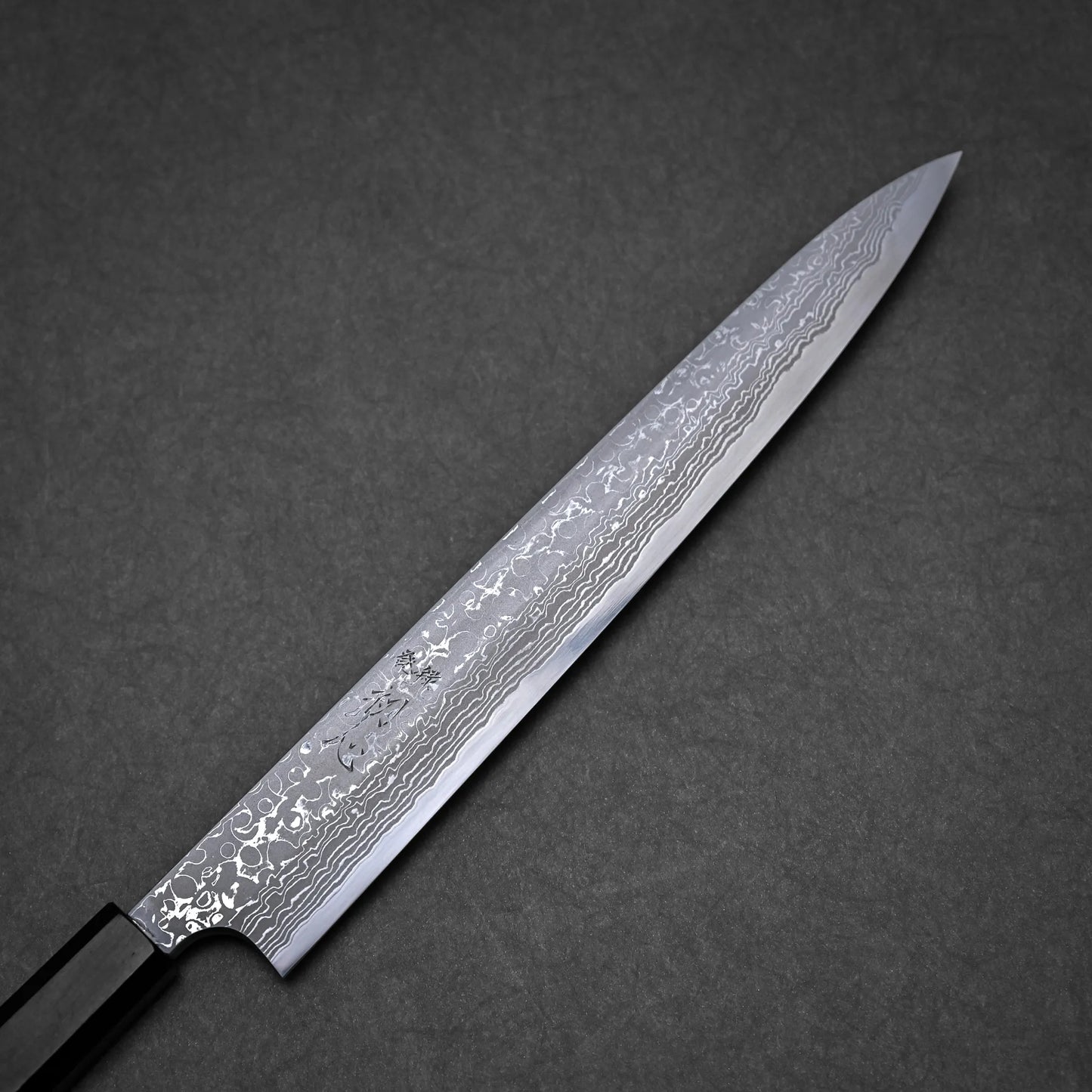 A Japanese-style damascus yanagiba with a long, narrow, and pointed blade featuring a visible Damascus steel pattern, set against a dark gray textured background.