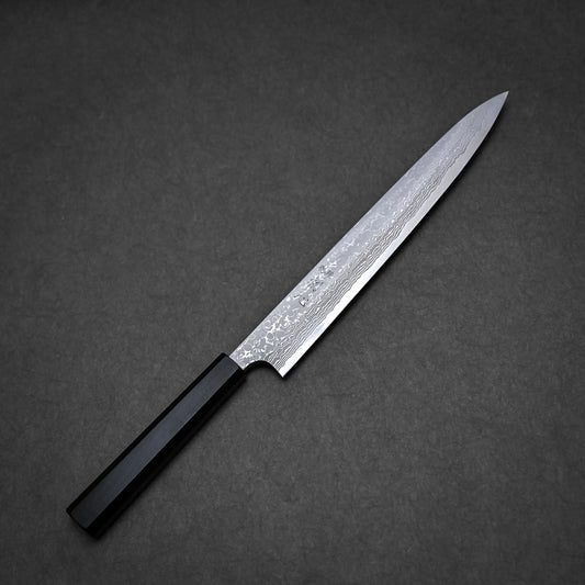 A Hatsukokoro x Yauji damascus aogami#2 yanagiba 270mm with a long, slender Damascus steel blade and a dark, angular handle is placed on a dark, textured surface.