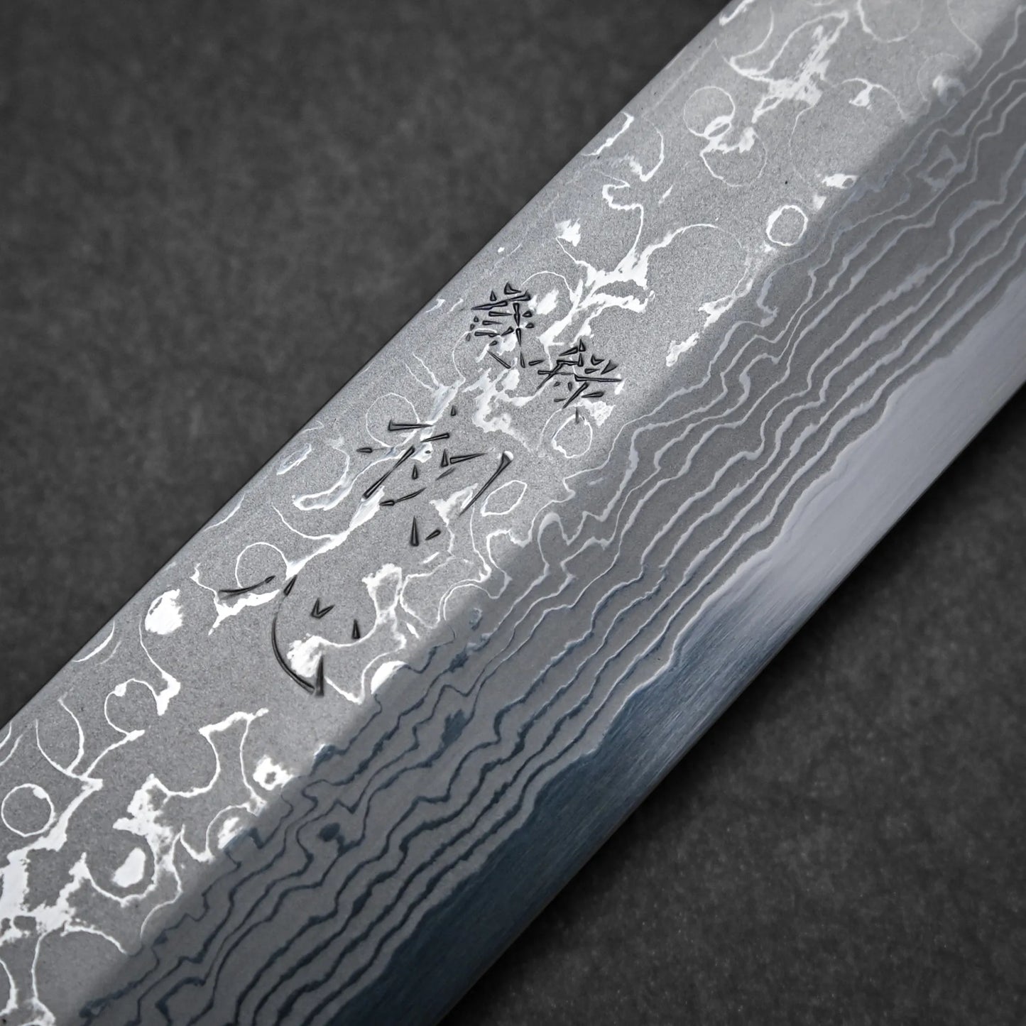Close-up of a Damascus sashimi knife blade showcasing intricate wavy patterns and Japanese characters engraved on the surface, set against a dark background.