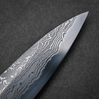 Close-up of the tip of a Damascus sashimi knife blade, showing distinctive wavy, layered patterns on the metal against a dark background.