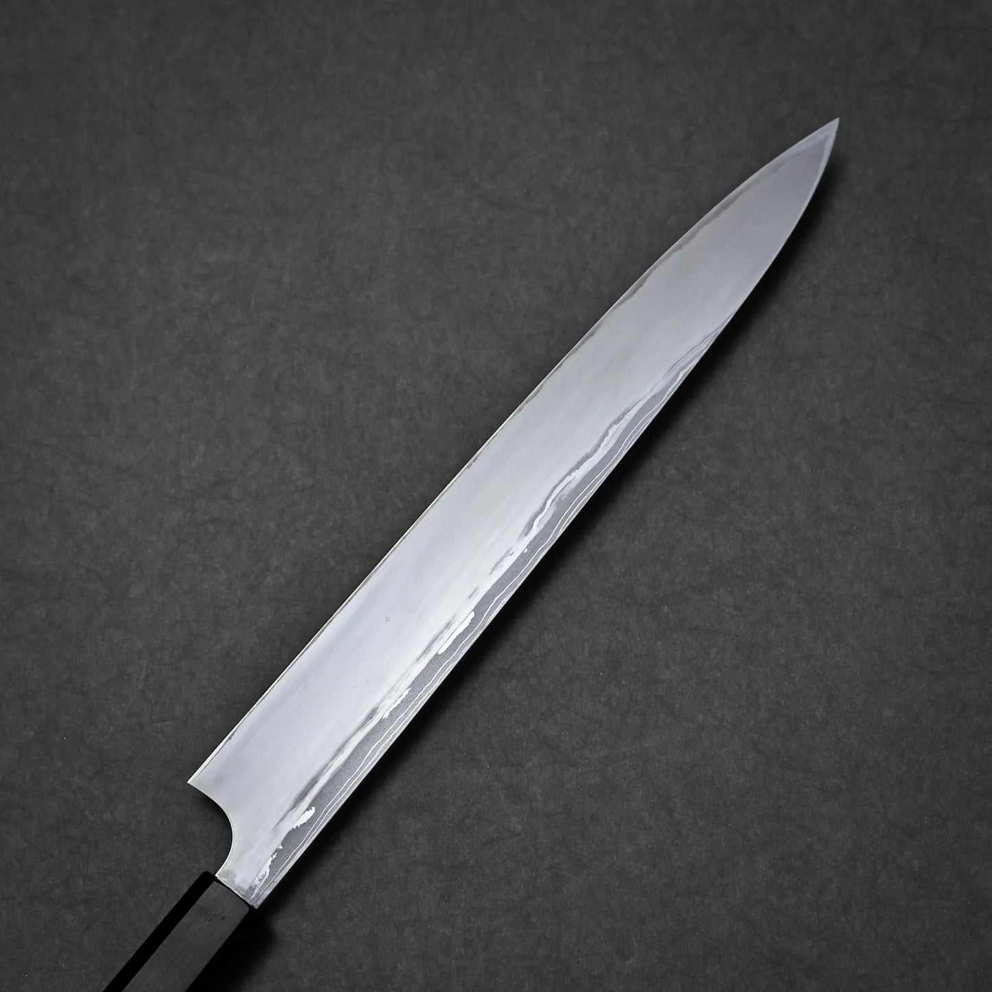 A close-up of a long, slender damascus yanagiba with a shiny, polished blade and a black handle, set against a dark, textured background.