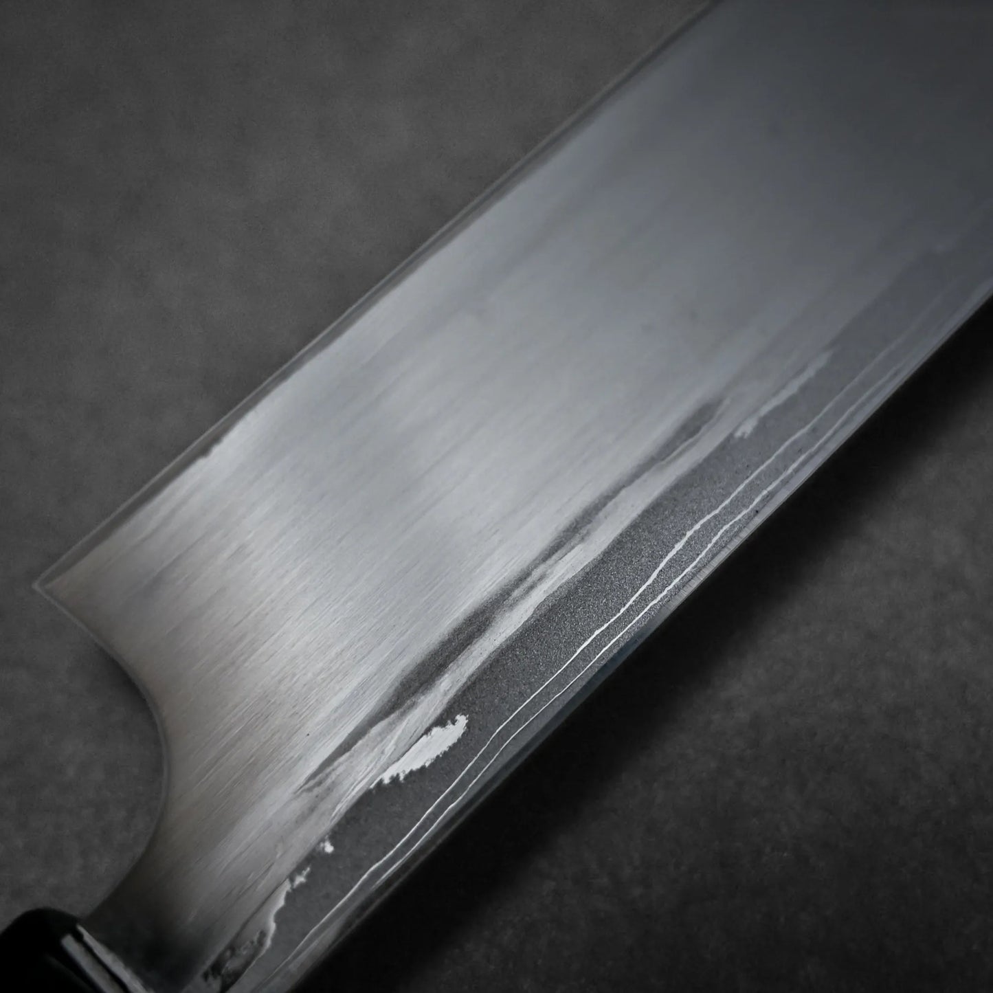 A close-up view of a damascus sushi knife blade showing a brushed metal finish and a distinct wavy line pattern around the uraoshi photographed against a dark, textured background.