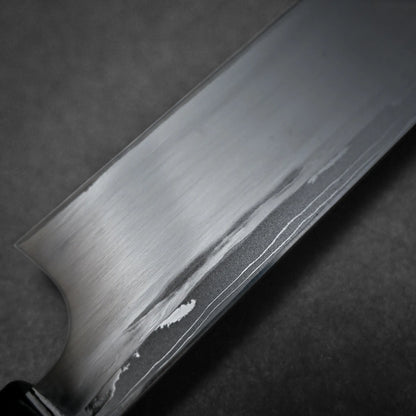 A close-up view of a damascus sushi knife blade showing a brushed metal finish and a distinct wavy line pattern around the uraoshi photographed against a dark, textured background.