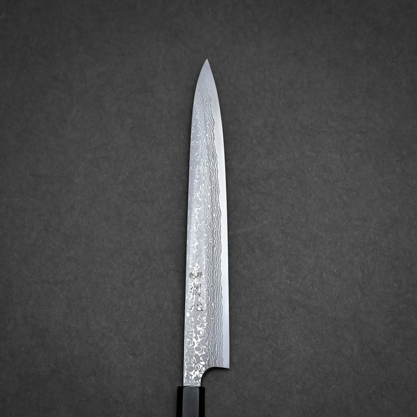A Japanese damascus yanagiba chefs knife with a patterned blade and black handle, positioned vertically on a dark, textured background. The blade displays intricate Damascus steel designs.