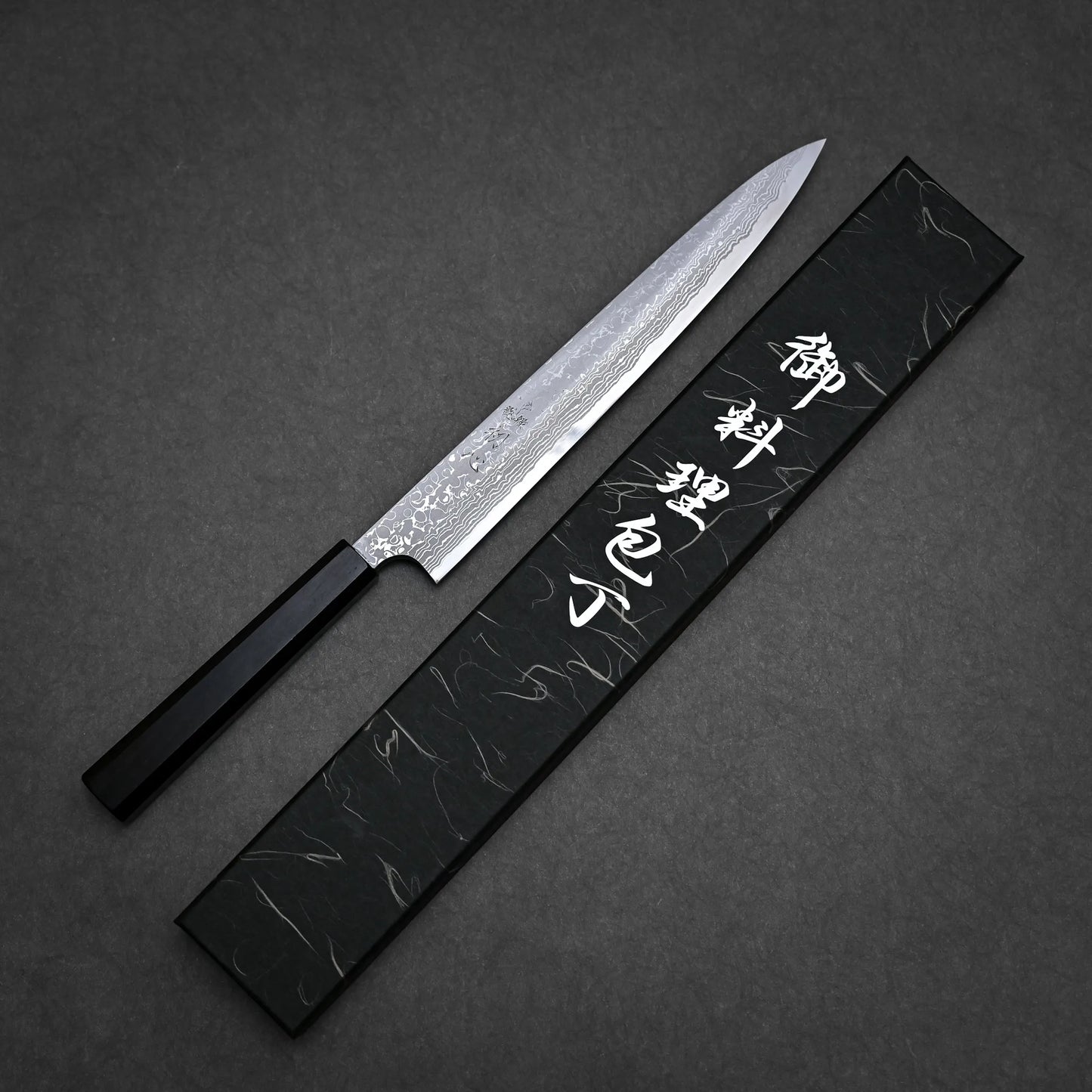 A Japanese-style damascus yanagiba with a patterned blade and black handle rests beside a black box adorned with white Japanese characters on a dark textured surface.
