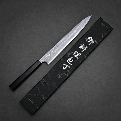 A Japanese-style damascus yanagiba with a patterned blade and black handle rests beside a black box adorned with white Japanese characters on a dark textured surface.