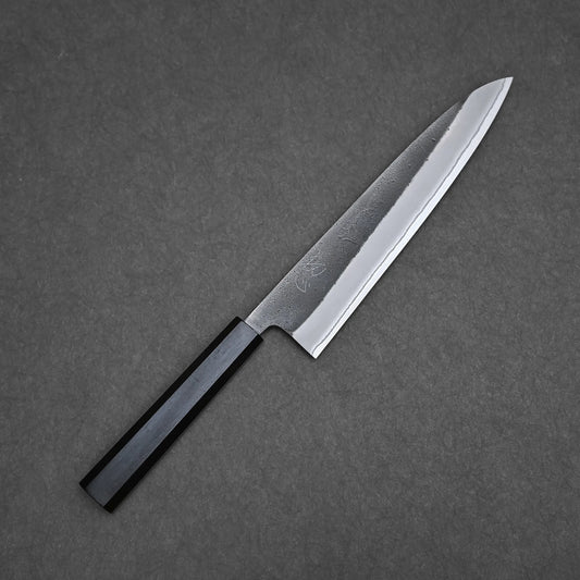 A Hatsukokoro x Yoshikane kurouchi nashiji shirogami#2 gyuto 210mm with a sharp, pointed blade and black octagonal handle rests on a dark, textured surface.